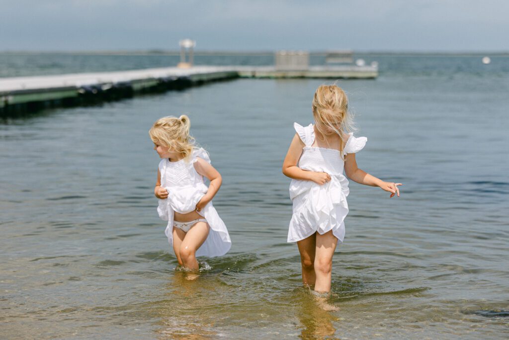 Two blonde girls in white dresses wading in the shallow water of the ocean tide. 