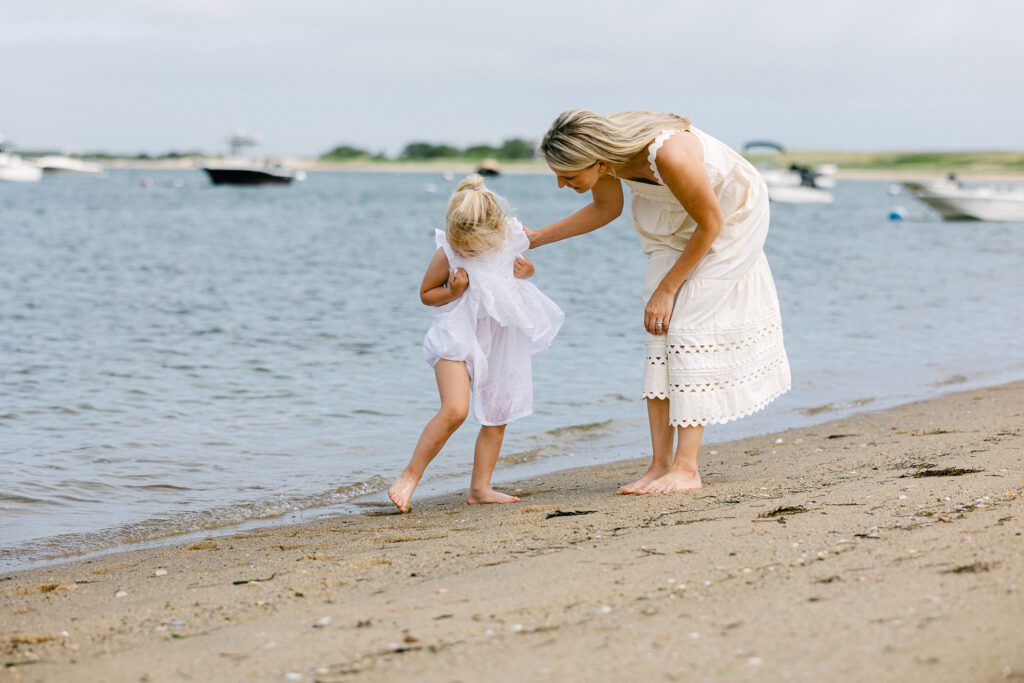 Blonde girl lifting her white dress up and looking at something on her leg, with mom bent over putting hand on her back to make sure she's OK. 