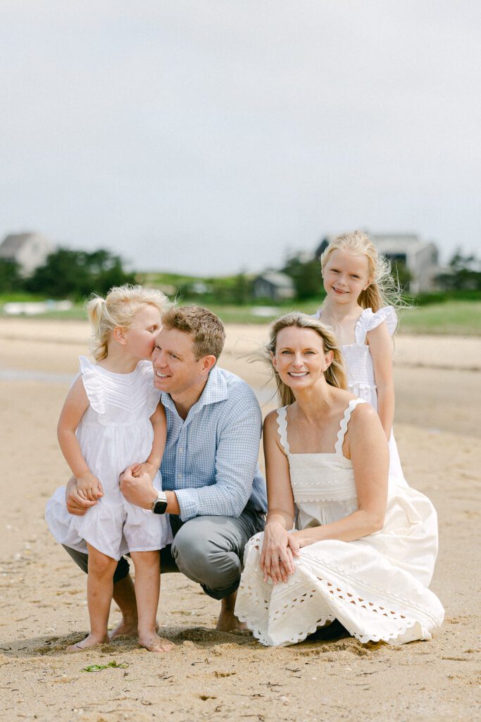 Family of four kneeling down on the beach. The youngest daughter stands in front of Dad and kisses his cheek while the oldest daughter stands behind Mom and smiles. 