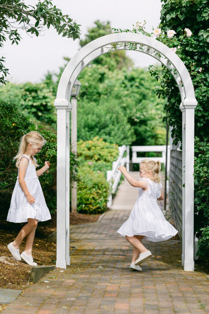 Blond girls in white dresses dancing in front of a white arch with greenery in the background.