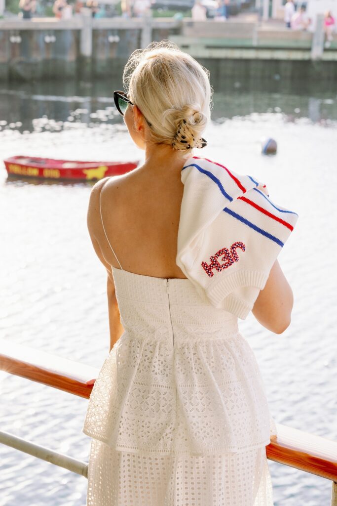 A blonde woman wearing a white eyelet dress with spaghetti straps is standing in front of a railing looking out onto the water where there is a small red boat. She has a red, white, and blue sweater with JEH embroidered along the neckline draped over her shoulder. 