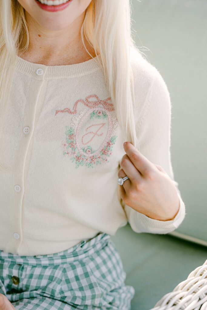 A blonde woman is holding her hair to the side to show off the floral embroidery surrounding a cursive J on an ivory cardigan is that buttoned all the way to the top. 