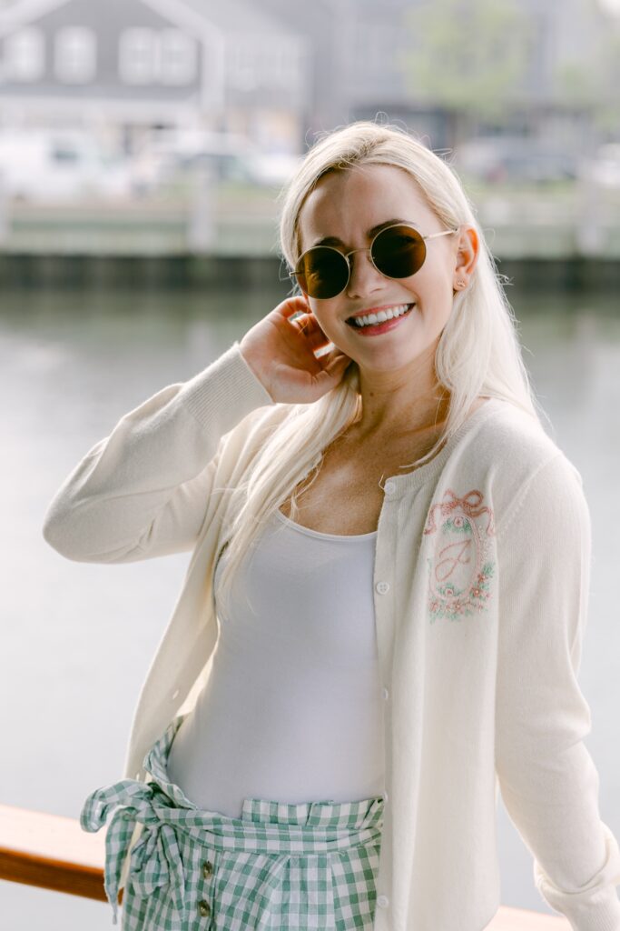 A blonde woman is putting her hair behind her ears, wearing round sunglasses and an ivory cardigan that ins unbuttoned featuring a floral embroidery surrounding a cursive J. She is smiling at the camera while leaning against a railing. 