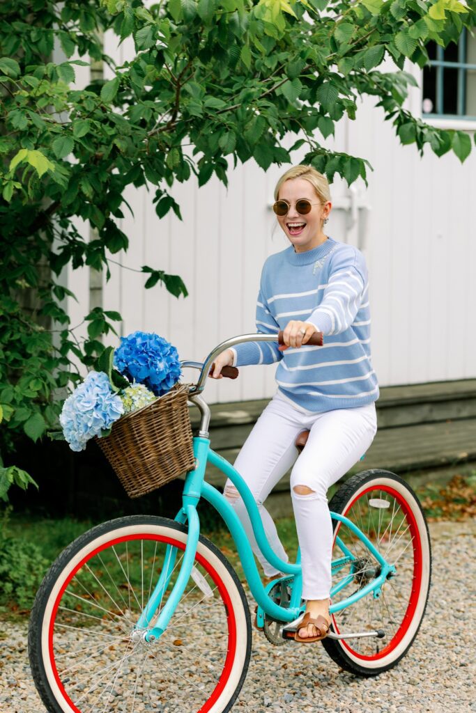 A blonde woman wearing round sunglasses and a sky blue and white striped sweater with ACK embroidered in crystals on the neckline is riding a turquoise bike down a gravel lane. The bike has a basket attached to the front filled with hydrangeas, and the woman is smiling. 