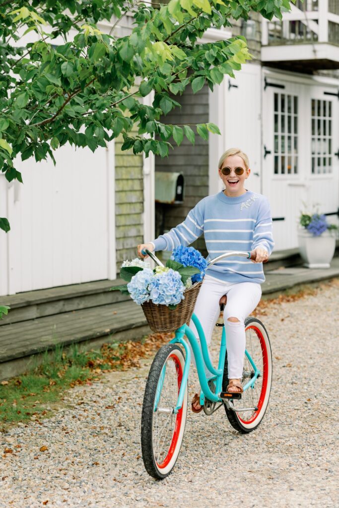 A blonde woman wearing round sunglasses and a sky blue and white striped sweater with ACK embroidered in crystals on the neckline is riding a turquoise bike down a gravel lane. The bike has a basket attached to the front filled with hydrangeas, and the woman is smiling. 