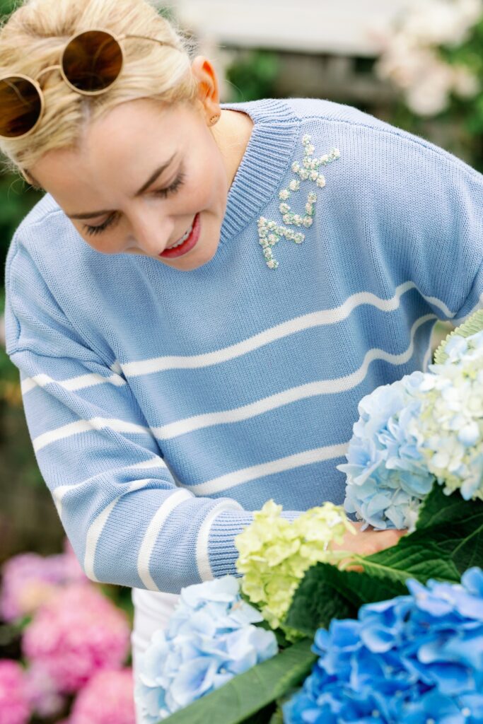 A blonde woman wearing a sky blue and white striped sweater with ACK embroidered in crystals on the neckline is leaning down looking at some hydrangeas, her hair pulled back and round sunglasses sitting atop her head. 