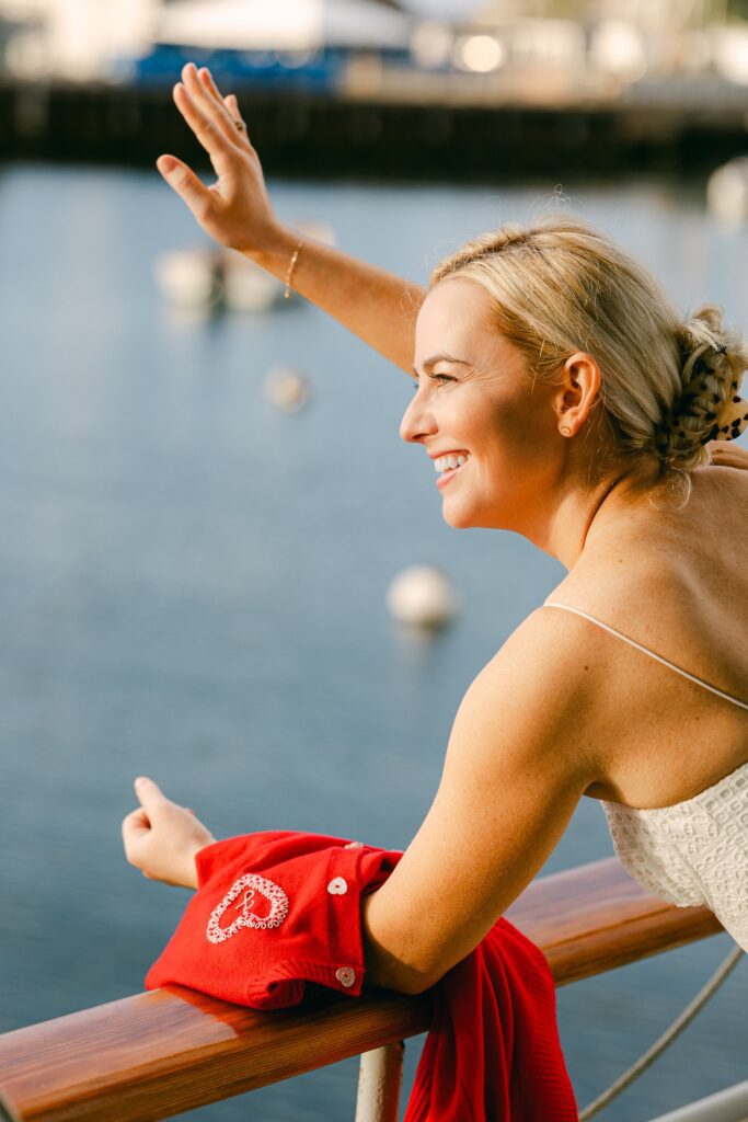 A blonde woman is smiling and waving while leaning against a railing whilst holding onto a red cardigan with a heart embroidered on it.