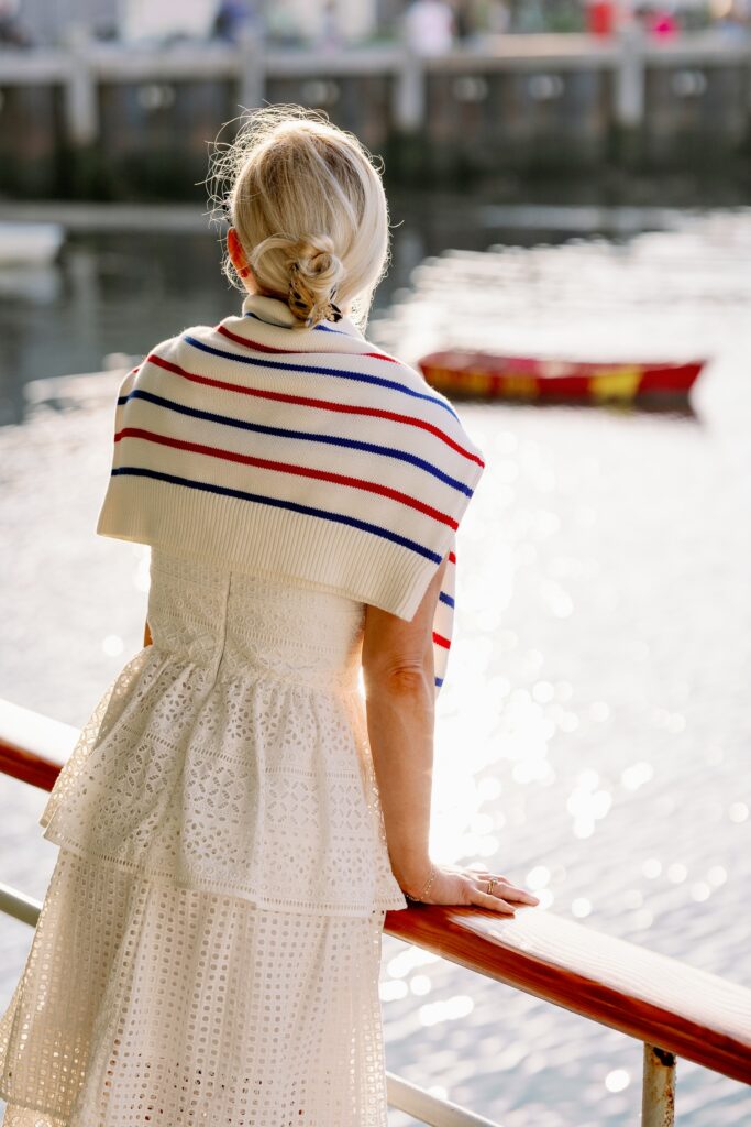 A blonde woman in a white eyelet dress has a blue and white striped sweater draped over her shoulders and is leaning on a railing looking over at the water. A small red boat is visible in the distance. 