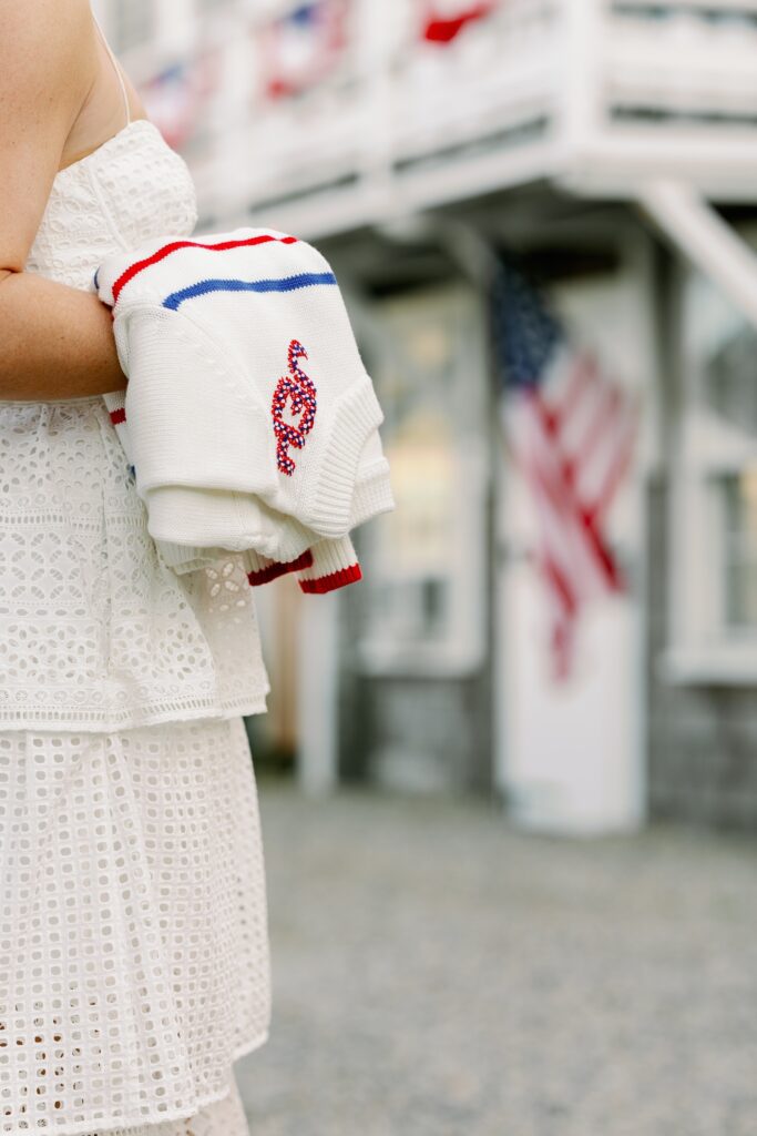 A woman in a white eyelet dress is standing and holding a red, white, and blue striped sweater hanging folded over her arm with visible embroidered letters. 