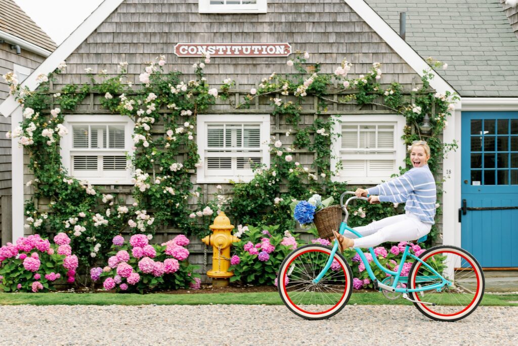 A blonde woman wearing a sky blue and white striped sweater and white jeans is riding a turquoise bike with a basket on the front holding hydrangeas. Both of her feet are lifted off the pedals and her legs are straightened I front of her - she is riding past a clapboard building covered in climbing roses with a few pink hydrangeas bushes blooming along the front. 