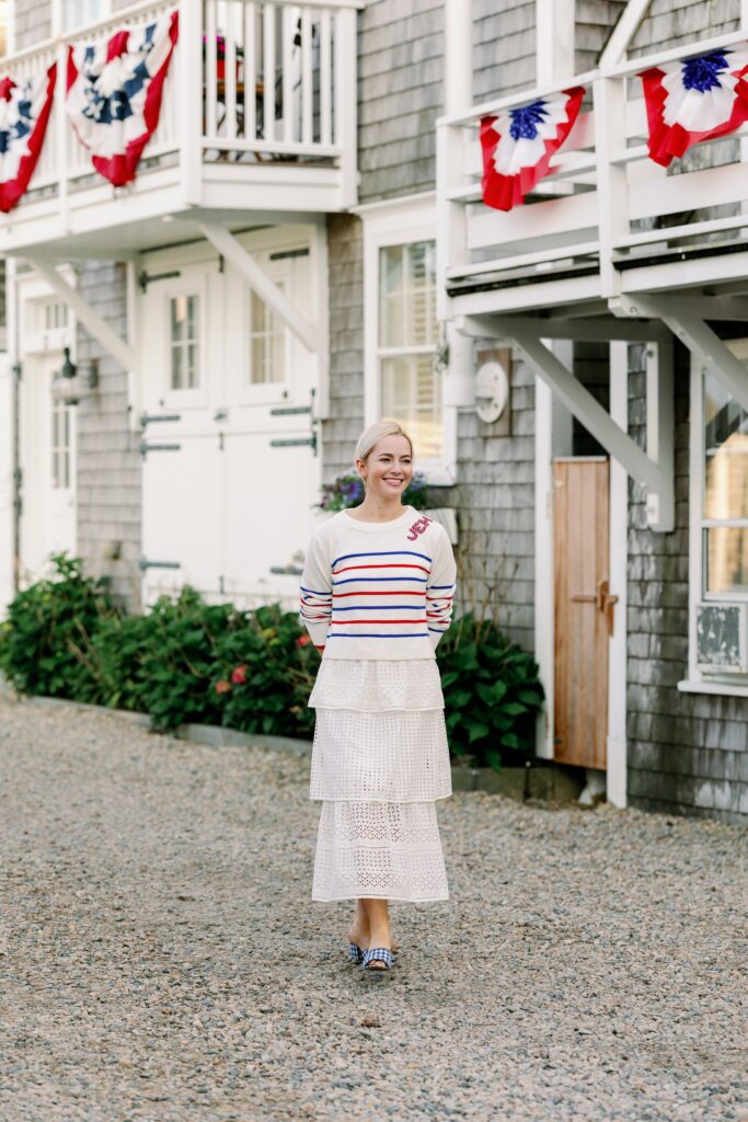 A woman is standing on a gravel road in front of a clapboard building with white balconies showcasing patriotic flags. She is smiling, wearing a red, white and blue striped sweater atop a white skirt. 