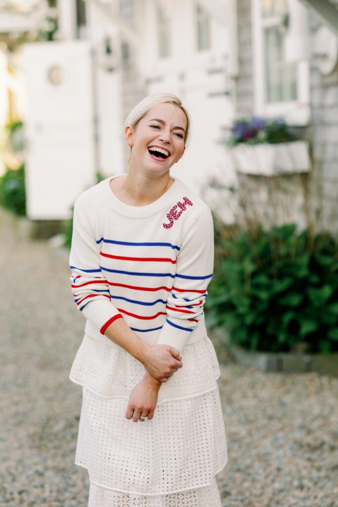 A woman is standing on a gravel road  smiling, wearing a red, white and blue striped sweater embroidered with JEH along the neckline atop a white skirt. 