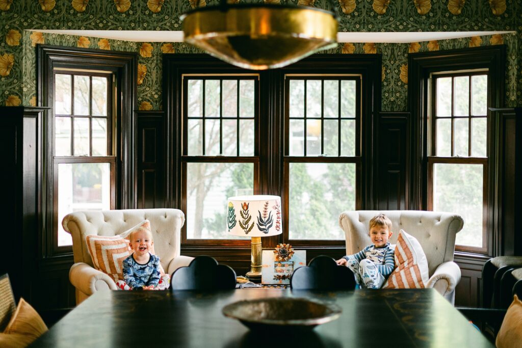 Blonde baby boy and brunette toddler boy sitting in matching cream chairs in front of bay windows flanking a table holding a gold lamp with a floral lampshade. Both in blue and white shark pajamas and smiling at the camera. 