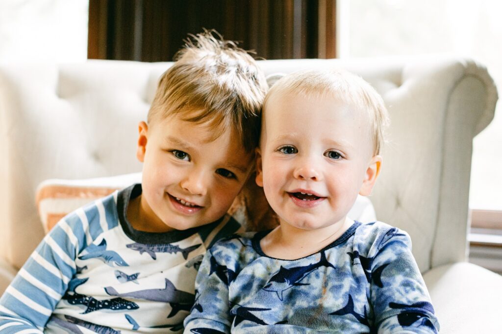 Brunette toddler boy and blonde baby boy are sitting side by side in complementing blue and white shark pajamas and smiling. 