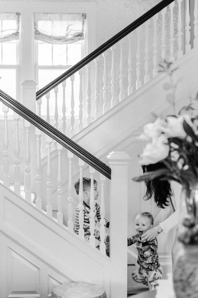 Toddler boy is sitting on the staircase while baby boy is standing at the bottom of the staircase holding onto the railing with a big smile on his face. 