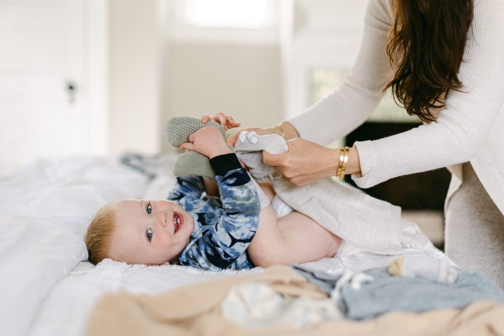 Blonde baby boy is laying on the bed holding his feet while mom is standing over him getting ready to put his pants on. 