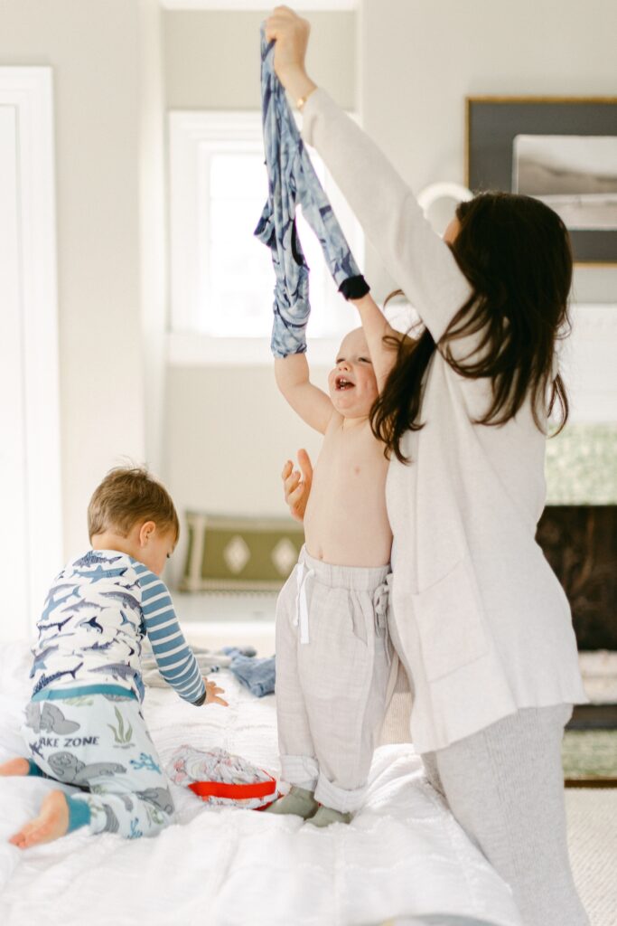 Blonde baby boy is standing on a bed with a big smile on his face and arms in the air as mom is standing behind him and pulling his pajama shirt off. Brunette toddler boy is kneeling on the bed in front of them looking at something laying on the bed. 