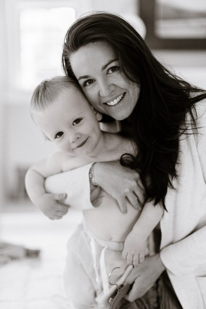 Mom is smiling with arm around baby boy who is shirtless with pants on and smiling as well. 