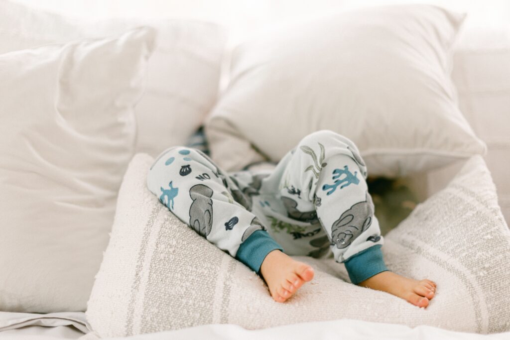 Toddler boy is laying under a handful of neutral pillows with only his knees down to his feet sticking out. 