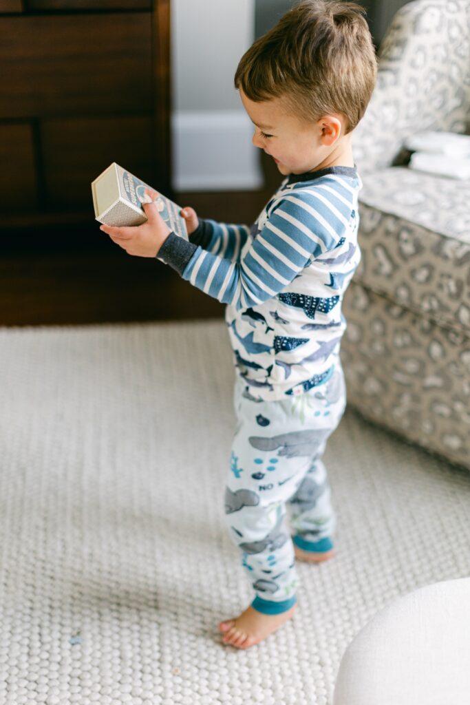 Brunette toddler boy in blue and white shark pajamas is standing up on an ivory rug while looking at a a little box containing a new toy. 