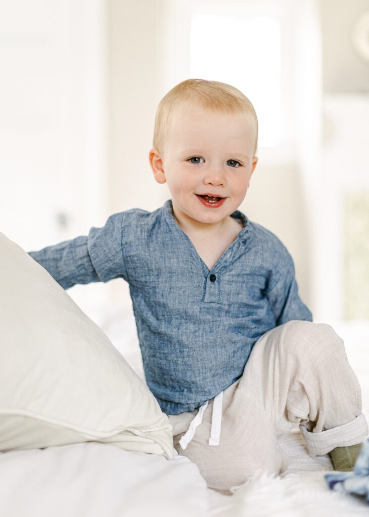 Blonde baby boy in a chambray shirt is kneeling on one knee, resting one hand on a pillow, and smiling. 