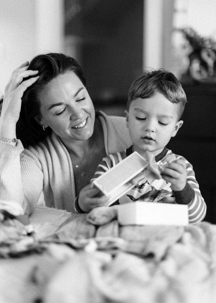 Mom is sitting with brunette toddler boy while he is opening a small box containing a new toy. 