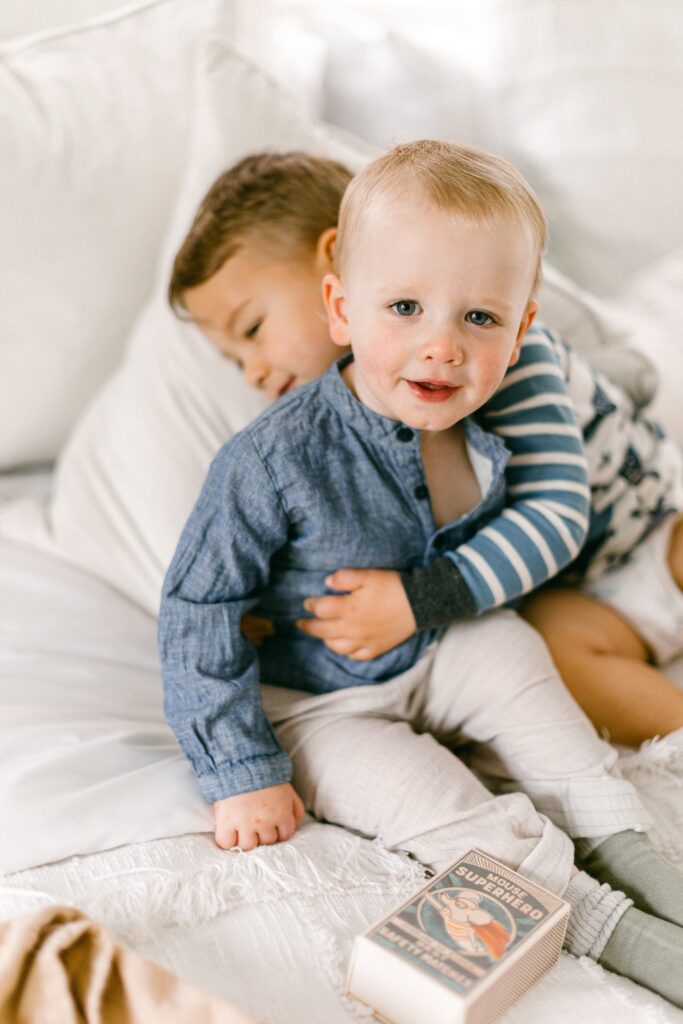 Blonde baby boy is sitting on the bed smiling while brunette brother has his around baby boy's waist while laying back on a pillow. 