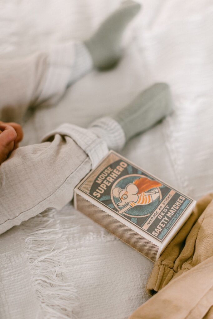 A small box of a Mouse Superhero toy is laying next to baby boy's leg's on the bed. 