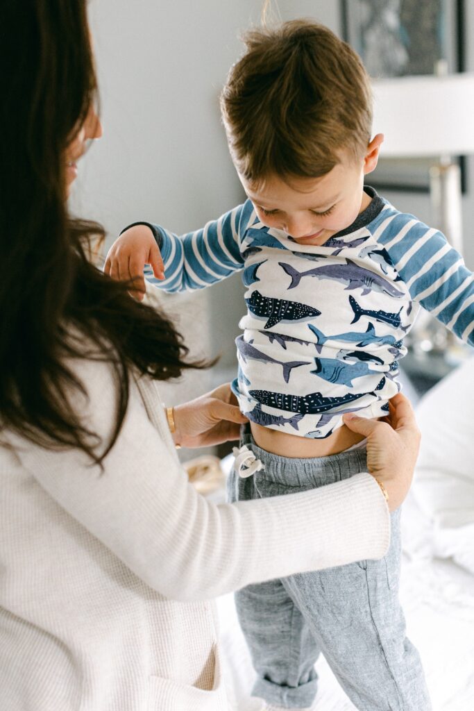 Toddler boy in nice pants and shark pajama shirt is getting dressed with help from mom who is standing in front of him and holding his waist. 