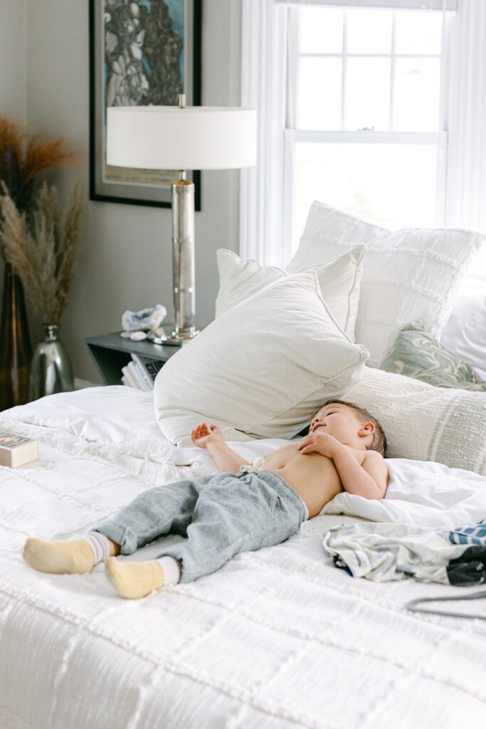 Brunette toddler boy is laying on a white bed shirtless with lots of pillows wearing nice pants and socks. 