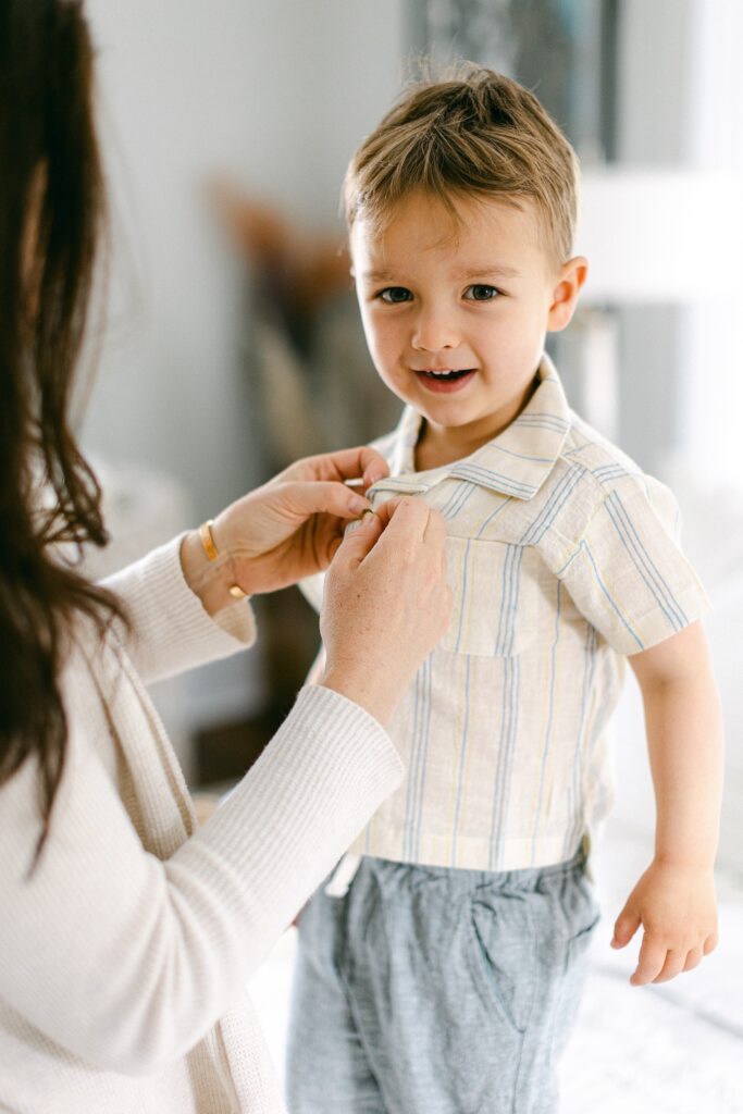 Smiling brunette toddler boy is getting help from mom buttoning the top button of his striped short-sleeve button down shirt. 