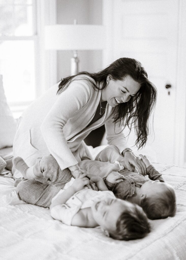 Two young boys laying side by side on their backs on the bed laughing as Mom leans over and tickles their bellies.