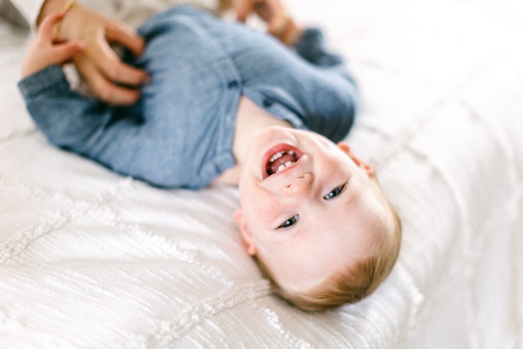 Blonde baby boy is showing off a toothy grin as he's laying on the bed with this head teetering close to the edge. 