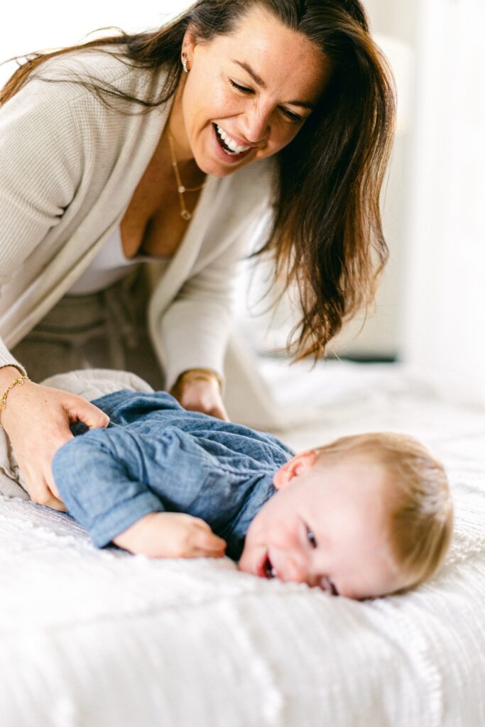 Blonde baby boy is laying on his belly on the bed laughing as a smiling mom  is squeezing his waist. 