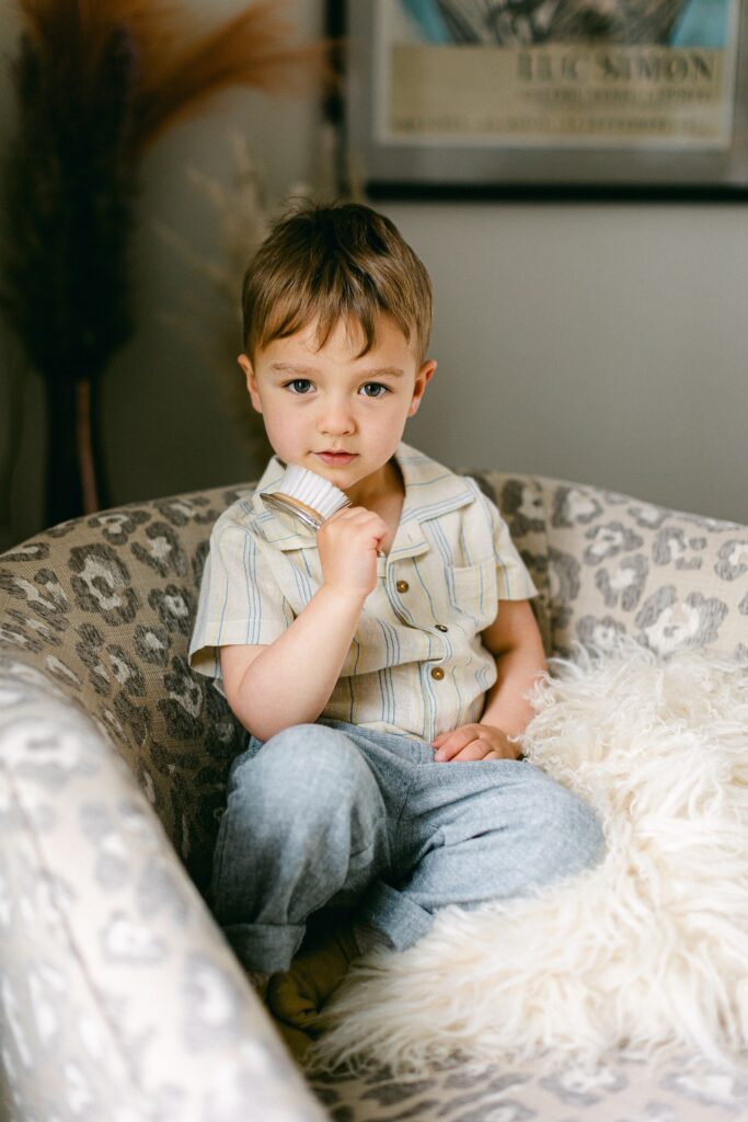 Brunette toddler boy is sitting on a neutral leopard print chair atop a fluffy pillow holding a silver baby's brush against his chin. 