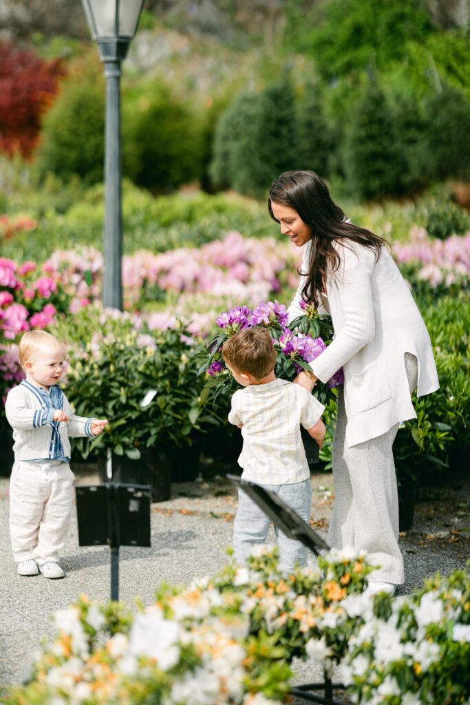 Brunette boy in a striped short-sleeve button-down shirt is being handed a flower pant with purple blooms to carry from mom. A blond toddler boy is standing in front and watching them while rows of flowers and shurbs are visible in the background. 