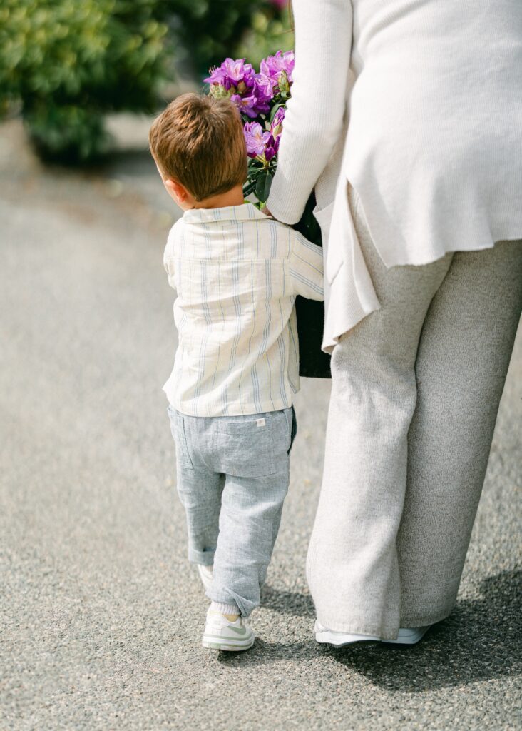 Brunette boy in a striped short-sleeve button-down shirt is being handed a flower pant with purple blooms to carry. 