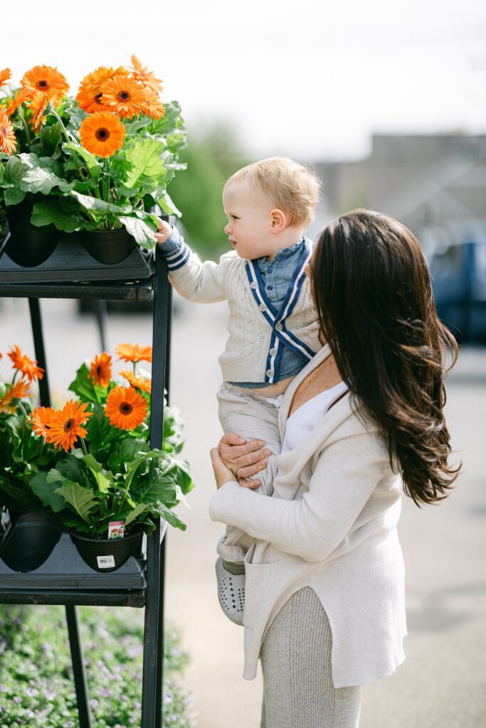 Brunette mom is holding up blonde baby boy wearing a chambray button-down and cable knit ivory cardigan. Baby is looking at a display of orange daises and mom is looking at the baby boy. 