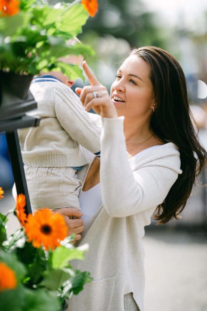 Brunette mom in a long ivory cardigan and white tank top is holding a child on her hip whose face is covered by a green leaf of the daisy flower display they are standing by, and mom is pointing up at something. 