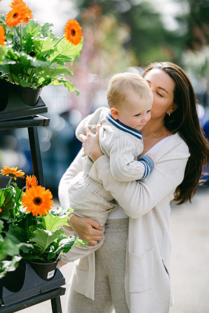 Mom with long brunette hair is holding blonde baby boy closely to her chest and kissing his cheek. 