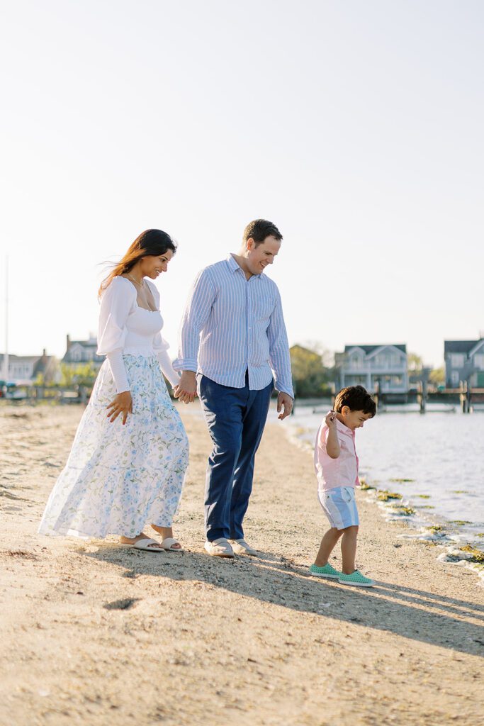 Brunette toddler boy is standing along the shoreline and is getting ready to throw a seashell into the water. Mom and dad are holding hands and smiling lovingly at him from behind.