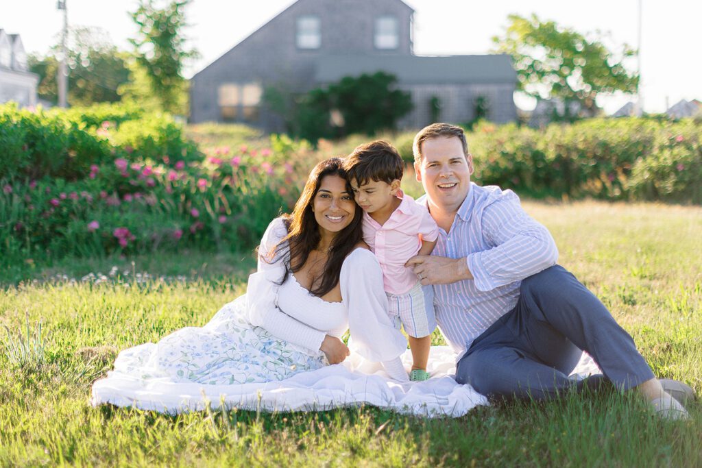 Mom and dad are sitting on a grassy patch with toddler on standing between them. Mom's skirt is splayed out and both mom and dad are smiling while toddler boy is looking down at something that caught his eye. 