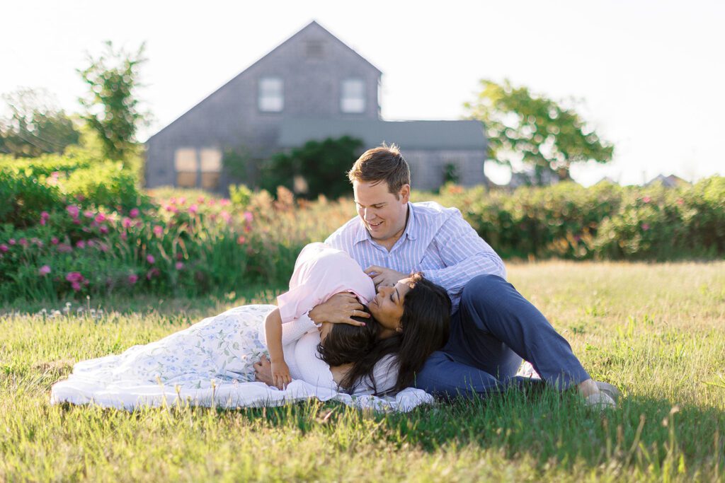 Dad is sitting on a grassy patch while mom is laying down on his leg and holding onto toddler's head as he is leaning over her shoulder resting his head on her chest. 