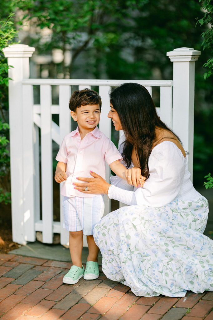 Brunette mom in a white top and floral skirt is kneeling down next to a brunette toddler boy in pink button down shirt and smiling at him. A white picket fence is right behind them.