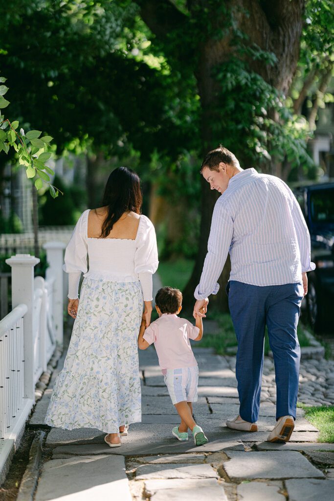 Brunette toddler boy in pink button down shirt and pastel striped shorts is walking in between mom and dad along a stone walkway holding both of their hands. All three are seen from behind. 