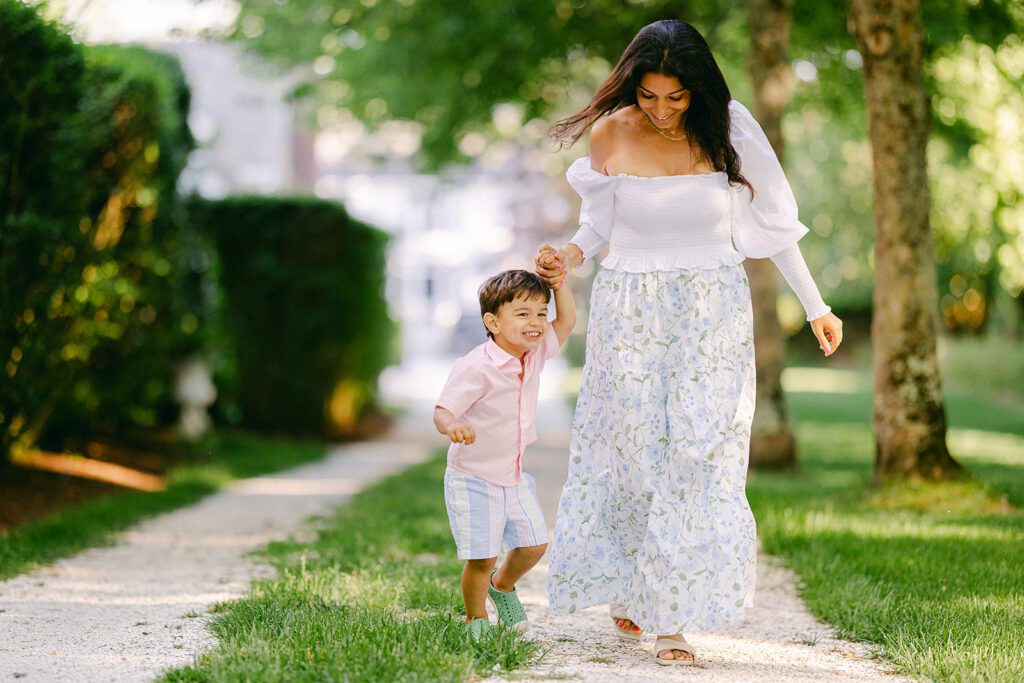 Mom in floral skirt is holding the hand of a brunette toddler boy smiling big  in a pale pink short sleeve button down and walking down a grass and shell lined path. 