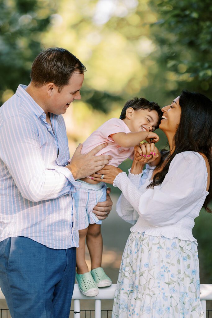 Dad is holding brunette toddler boy on top of a white fence - the toddler boy is laughing and leaning into a smiling mom in a white blouse and floral skirt standing beside him. 