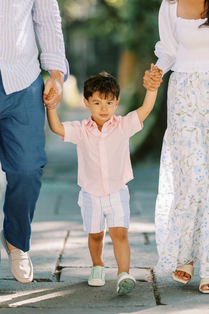 Brunette toddler boy in pink button down shirt and pastel striped shorts is walking in between mom and dad holding both of their hands wearing a serious expression. 