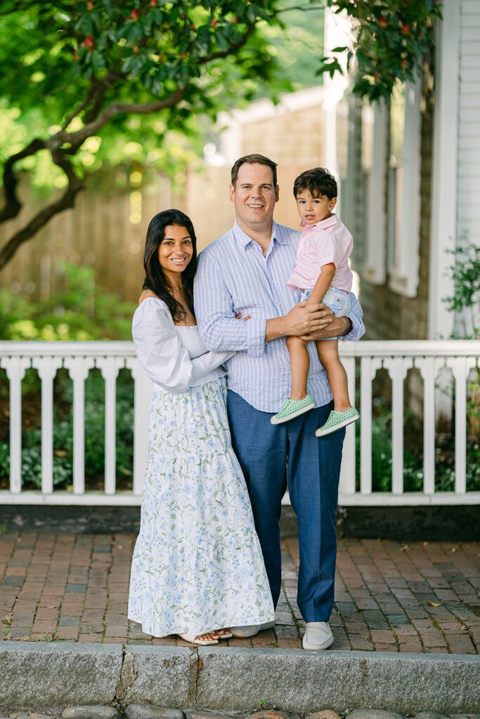 Mom, dad, and brunette toddler boy are smiling at the camera, all dressed in shades of blue and green, with a pop of pink from the toddler's shirt, on a brick sidewalk with a white fence behind them. 