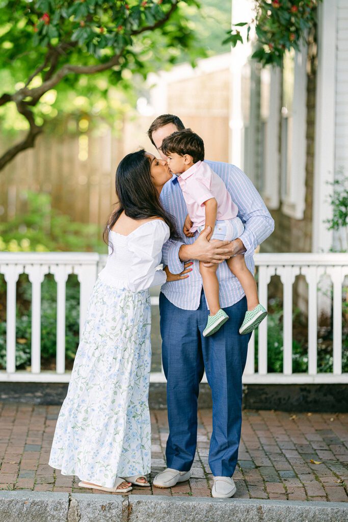 Dad in blue slacks is holding brunette toddler boy who is leaning over to kiss brunette mom. They are on a brick sidewalk with a white fence behind them. 